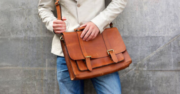 Eine Person trägt eine elegante, braune Unisex-Businesstasche aus Leder vor einer Betonwand.